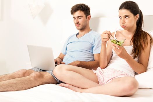 Addicted young man on bed with a laptop while the woman looks angry