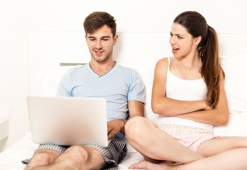 Addicted young man on bed with a laptop while the woman looks angry