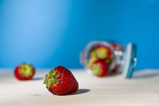 Close up of a strawberry and a glass jar full of strawberries lying down on a table with blue background