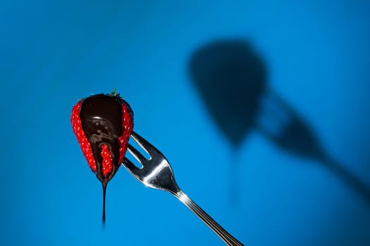 Close up of a strawberry suspended from a fork and its shadow on background with liquid chocolate above
