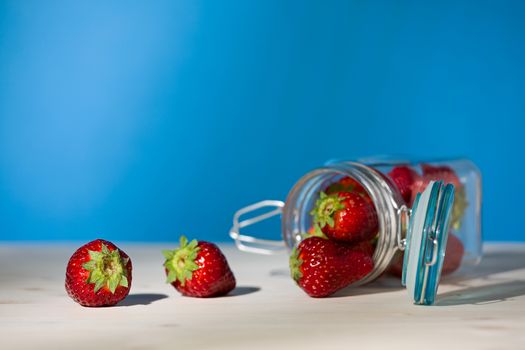 Strawberries and a glass jar full of strawberries lying down on a table with blue background