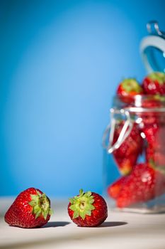 Strawberries on a table with blue background and a glass jar full of strawberries on the background