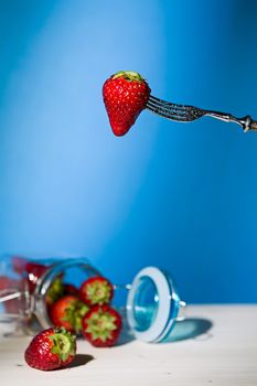 Strawberry suspended from a fork and under a jar of strawberries with blue background