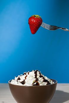 Strawberry suspended from a fork over a cup of whipped cream and chocolate with blue background