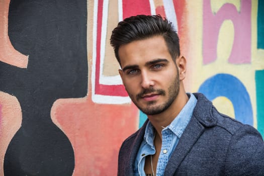 Attractive young man standing against colorful graffiti wall, looking at camera