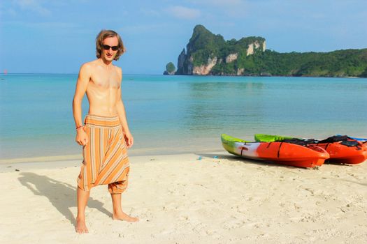 a young topless man standing in front of a row of Kayaks canoes boats on the PhiPhi Don beach in Thailand