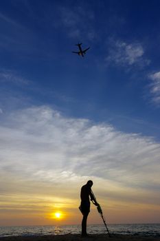 a treasure hunter with Metal detector on sunset on the beach, with a plan in the sky.