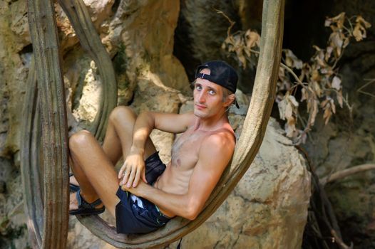 a shirtless young man relaxing and leaning against tree or a liana in front of cave