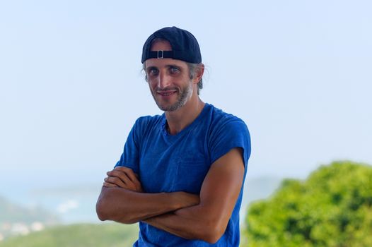 a happy Young Bearded Man sitting on the stone railings in city park wearing in blank blue t-shirt and black Cap