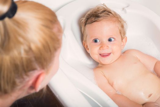 Little pretty wet baby boy in bath room sitting and smiling with mother's hand on white background, horizontal picture.