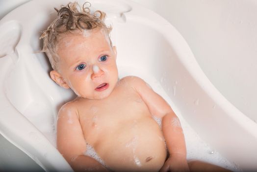 Little pretty wet baby boy in bath room sitting and sad on white background, horizontal picture.