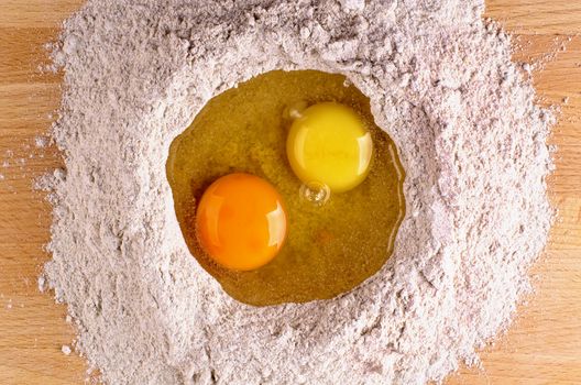 Two Light Yellow and Dark Yellow Egg Yolks into Heap of Whole Wheat Flour closeup on Wooden Table