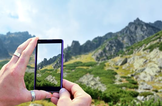 View over the mobile phone display during taking a picture of landscape in nature. Holding the mobile phone in hands and taking a photo. Focused on mobile phone screen. 