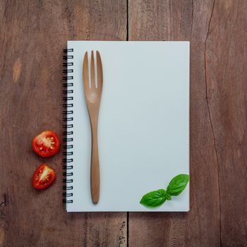 Foods background and Food menu design . Notebook background for recipes and shopping list. Notebook ,sweet basil ,cherry tomatoes slice and fork on dark shabby wooden background flat lay .