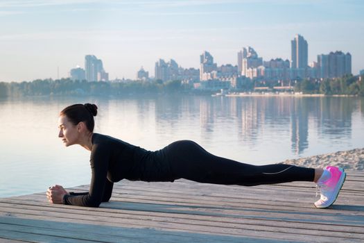 Young pretty fitness woman exercises on pier during morning sport training workout