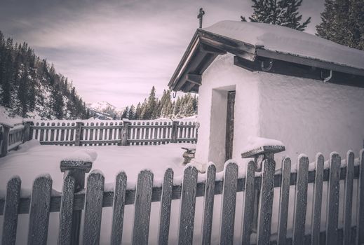 Winter scenery with a small, rustic chapel covered with snow, surrounded by a wooden fence and snowy trees. Image captured in Ehrwald, Austria.