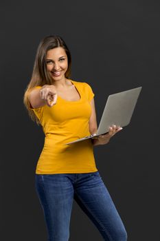 Beautiful woman in front of a black wall holding a laptop and pointing 