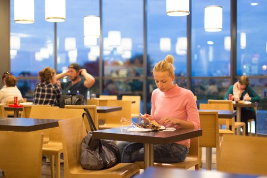 Young female traveler eating pizza at airport restaurant while waiting for late night flight departure.