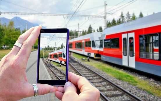 View over the mobile phone display during taking a picture of train at railway station. Holding the mobile phone in hands and taking a photo. Focused on mobile phone screen.