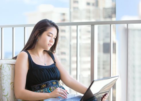 Beautiful teen girl sitting on balcony on highrise building in large city using laptop computer