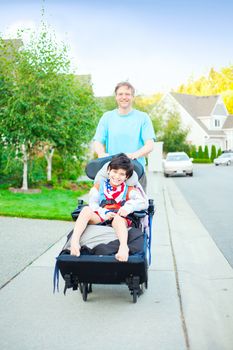 Caucasian father pushing disabled biracial  ten year old son in wheelchair outdoors along sidewalk. Child has cerebral palsy.