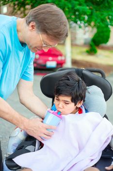 Father holding water cup, helping disabled ten year old son in wheelchair drink from straw