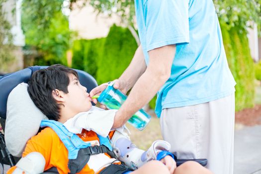 Father holding water cup, helping disabled ten year old son in wheelchair drink from straw