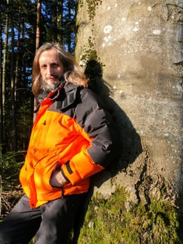 a bearded man in a orange jacket standing in front of a tree with morning sunlight