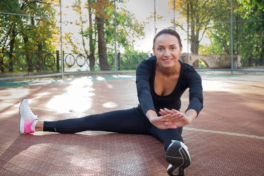 Young pretty fitness woman does stretching exercises during sport training workout on playground outdoor