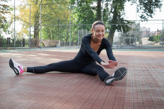 Young pretty fitness woman does stretching exercises during sport training workout on playground outdoor