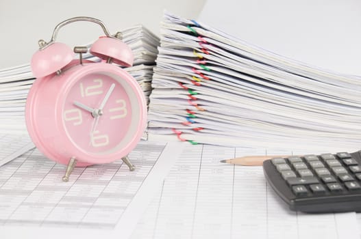 Brown pencil and old pink alarm clock on finance account have blur calculator and pile overload paperwork of report and receipt with colorful paperclip as background.