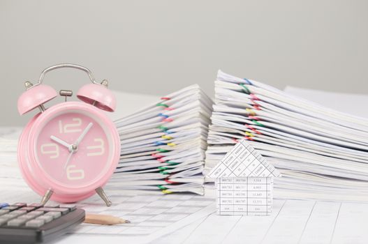 House on finance account have blur old pink alarm clock and calculator with pencil and pile overload paperwork of report and receipt with colorful paperclip as background.