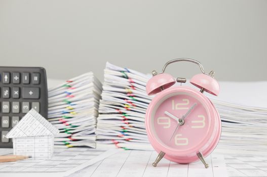 Old pink alarm clock on finance account have blur house and calculator with pencil and pile overload paperwork of report and receipt with colorful paperclip as background.