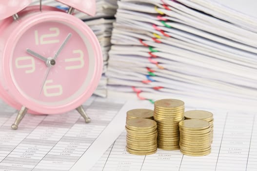 Pile of gold coin on finance account have blur old pink alarm clock and pile overload paperwork of report and receipt with colorful paperclip as background.