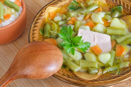 Soup of pork with vegetables and rice, a ceramic bowl and a wooden spoon. Stands on table, close-up.