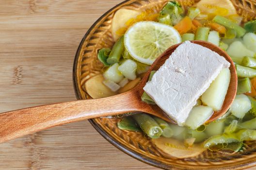 Soup of pork with vegetables and lemon, a ceramic bowl and a wooden spoon. Stands on table, closeup.