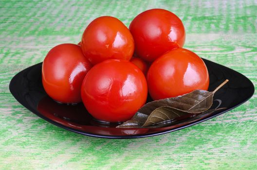 Pickled red tomatoes on a black plate and green old wooden background.