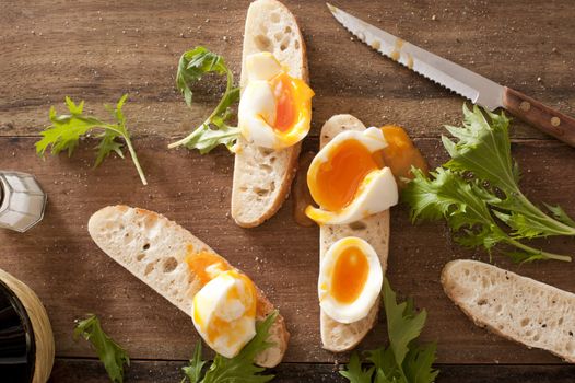 Egg soldiers made from soft boiled egg sliced onto freshly baked slices of baguette with strewn rocket leaves on the surface of a wooden table for a tasty finger snack