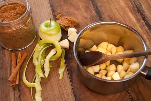 Preparing homemade apple and cinnamon sauce with fresh peeled and sliced green apples in a pot alongside stick cinnamon and ground spice on a wood table