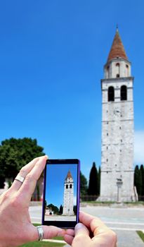 View over the mobile phone display during taking a picture of Basilica of Aquileia in Italy. Holding the mobile phone in hands and taking a photo of Basilica of Aquileia. Focused on mobile phone screen.