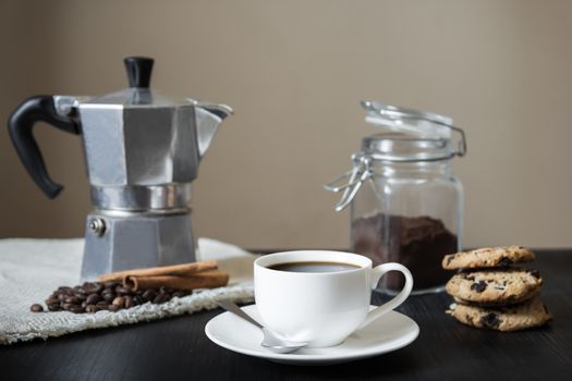 Front view of cup of black coffee, italian coffee pot on flax table-napkin and chocolate chip cookies on black vintage wood table indoors in natural light