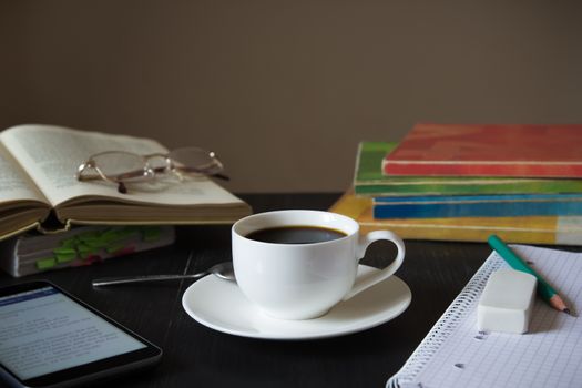 Black wood table with cup of coffee, textbooks, smartphone, reading glasses and notebook