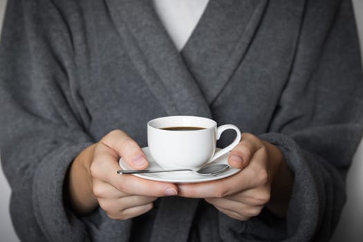 Hands of female person in bathrobe holding cup of morning coffee in front of white wall