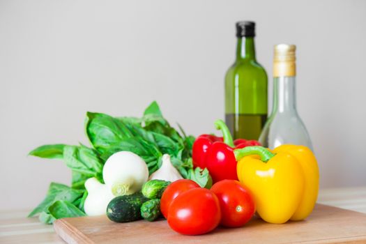 Fresh vegetables, basil, olive oil and vinegar on wood kitchen table in front of white wall