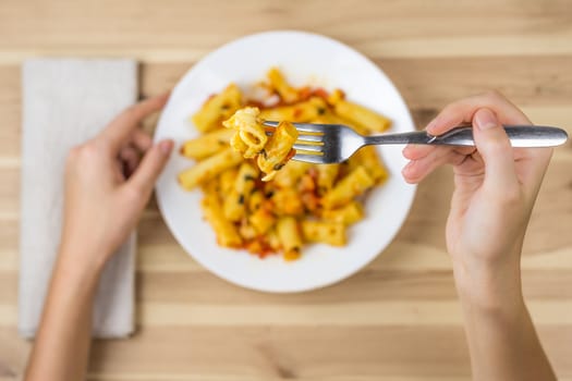 Cooked tortiglioni pasta on fork close up. Female hand holding fork above the plate of baked pasta.