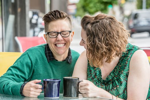 Cute young lesbian couple joking while seated outside at table with coffee mugs