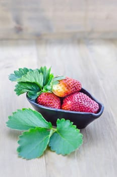 fresh strawberry fruits in black bowl with green leaves over wooden background