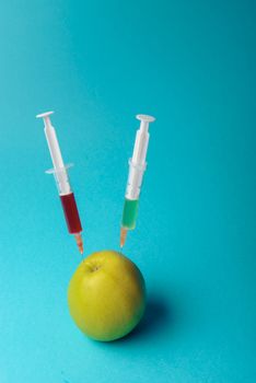 Chemical additives in food or genetically modified fruit concept. Green apple with syringes of chemicals. Isolated on blue background.