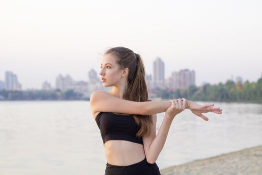 Young pretty slim fitness sporty woman stretches preparing for training workout exercises outdoor at river coast in the morning