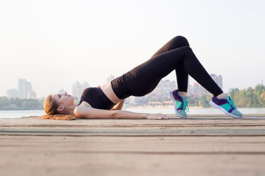 Young pretty slim fitness sporty woman doing butt-sculpting exercises during training workout outdoor at river coast in the morning